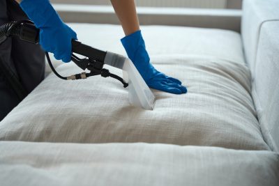 Worker standing in the bedroom near the sofa, holding vacuum cleaner and cleaning seat cushions