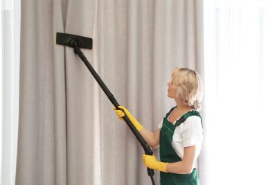 Female janitor removing dust from curtain with steam cleaner indoors